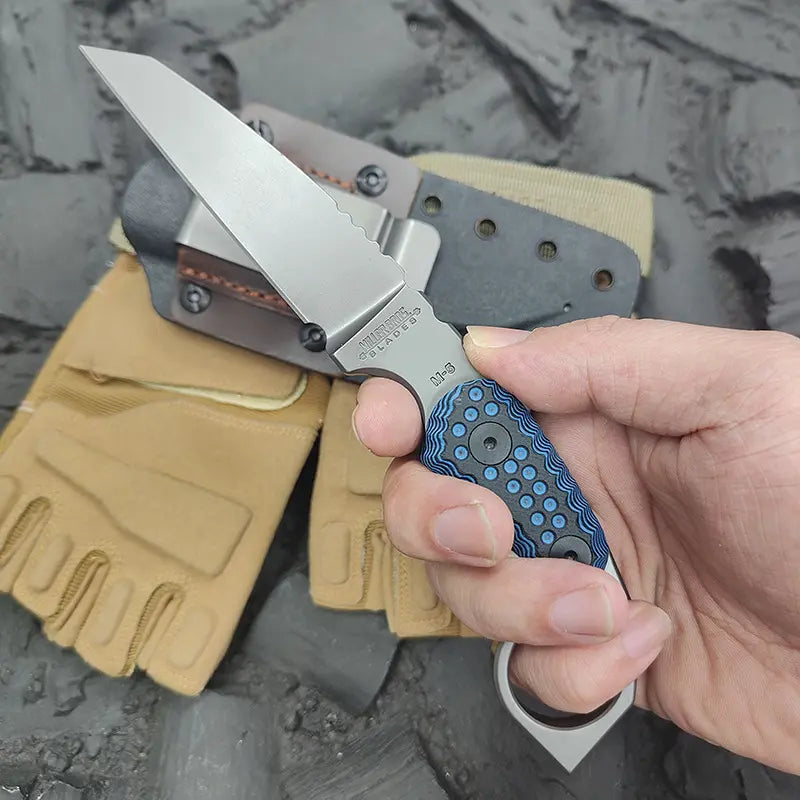 Hand holding a folding knife with a textured blue handle, blurred gloves and stone background