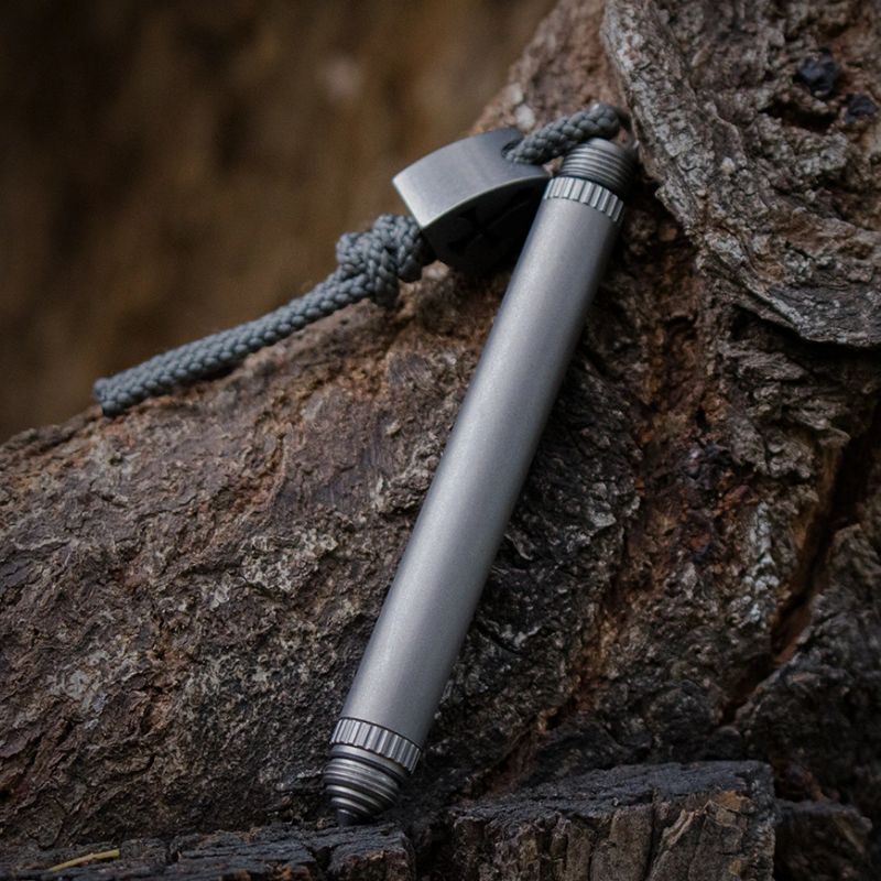A titanium Mantis Pen 4-in-1 multi-tool with a micro knife and a Tungsten Steel attack head, placed on a rock, with a paracord bracelet attached to it.